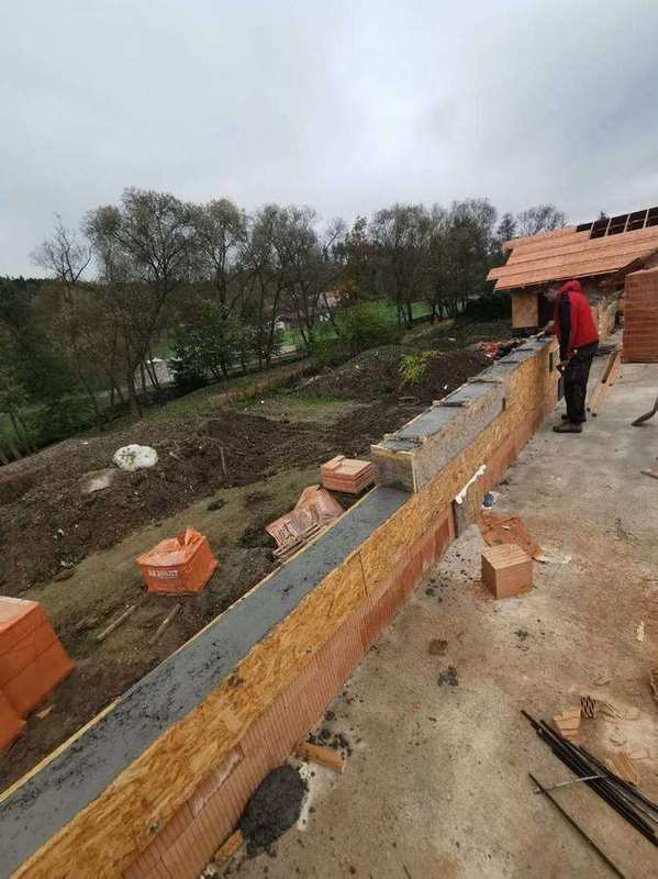Pracovník hlazí čerstvý beton věnce na obvodové stěně z Heluz cihel – pohled podél zdiva na podzimní krajinu