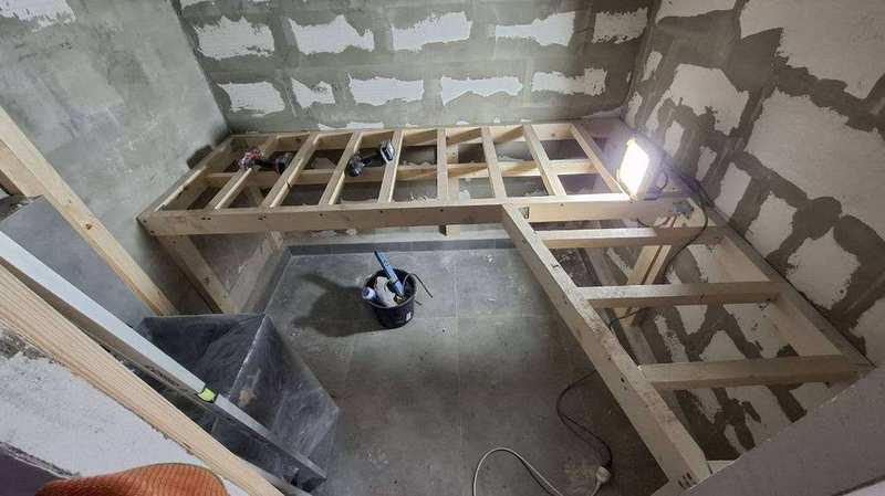 Interiér sauny s hotovým smrkovým obložením stěn a roštem lavic – stavební náčiní na lavici, LED páska u podlahy, pohled na třístupňové sezení