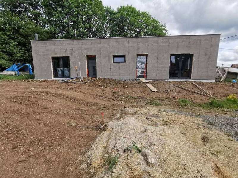 Přední pohled na dokončenou hrubou stavbu bungalovu – fasáda s hrubou omítkou, čtyři okenní otvory a terénní úpravy v průběhu