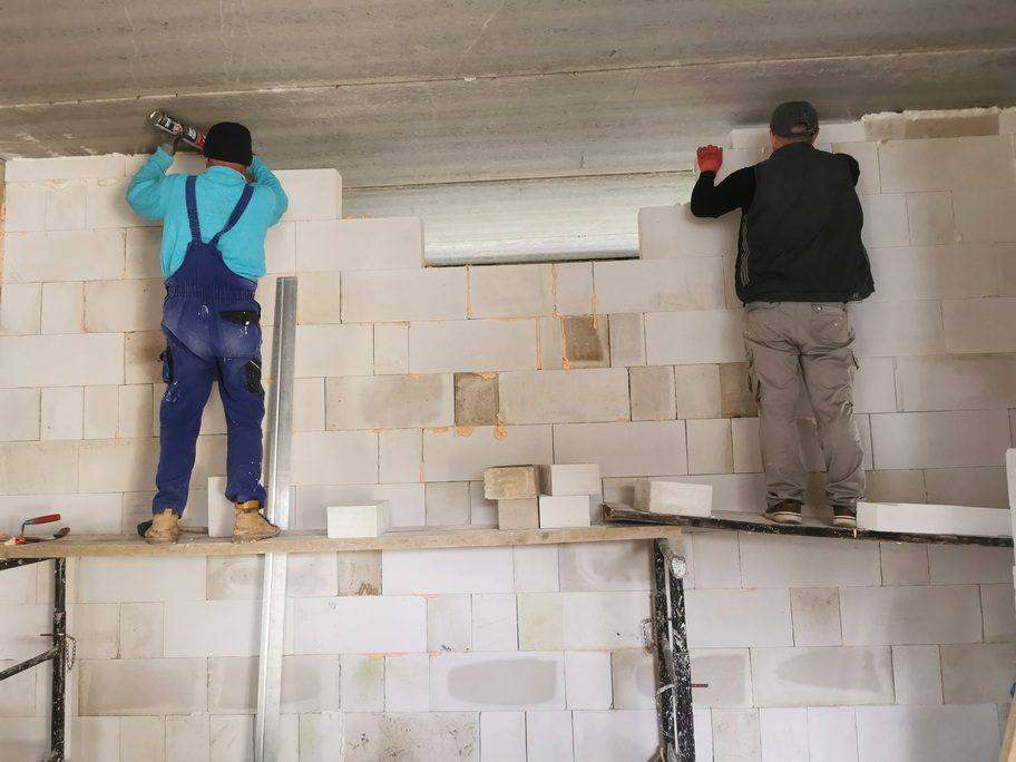Dva zedníci aplikují PUR pěnu pod stropní panel na vrcholu obvodové zdi – dokončování poslední řady